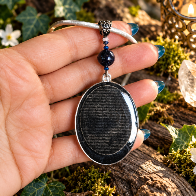 Dos du médaillon ovale du collier Le Miroir aux Fées, avec son revers sombre bordé de métal argenté, présenté dans un décor naturel.