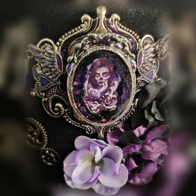 Médaillon émaillé du chapeau Mina – Portrait gothique inspiré de Dracula Zoom sur le médaillon émaillé à la main du chapeau steampunk Mina représentant Mina vampire, fleurs violettes, lilas et noires