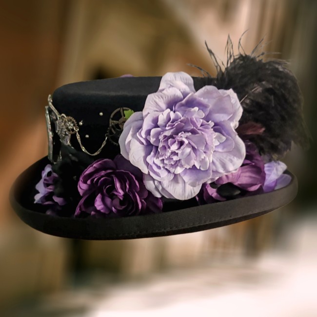 Chapeau Mina vue latérale – Fleurs textiles, plumes et mécanique steampunk Chapeau steampunk gothique Mina vue de gauche avec fleurs en tissu, engrenages métalliques et plumes d’autruche noires