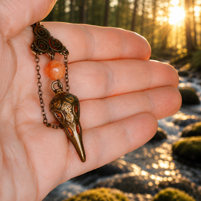 Gros plan sur Le Veilleur des Runes, collection Hrafnseiðr. Ce bijou artisanal inspiré de la mythologie nordique dévoile un pendentif corbeau finement gravé, des perles orange lumineuses et des chaînes bronze délicates dans une ambiance de forêt scandinav