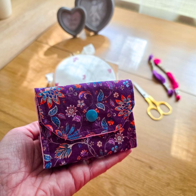 Gros plan sur la prise en main de la mini poubelle à fils nomade et pliante modèle Cachemire en tissu violet aux motifs floraux et cachemire, fermée par bouton-pression pour se glisser dans une trousse de brodeuse ou pochette à projet, création artisanale