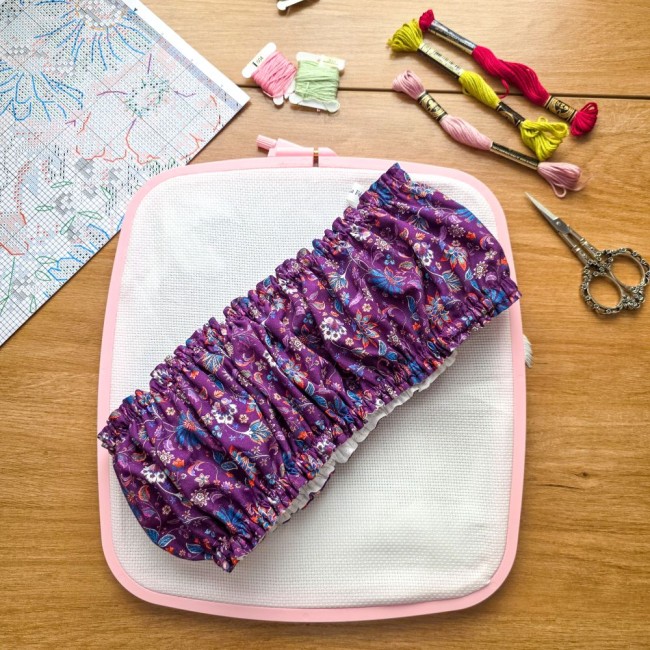 Vue de dessus d'un grime guard housse protège-toile modèle Cachemire en tissu violet aux motifs floraux et cachemire multicolores, posé en travers d'un tambour rectangulaire Nurge rose avec ciseaux brodeuse, échevettes de fils et grille de broderie, créat