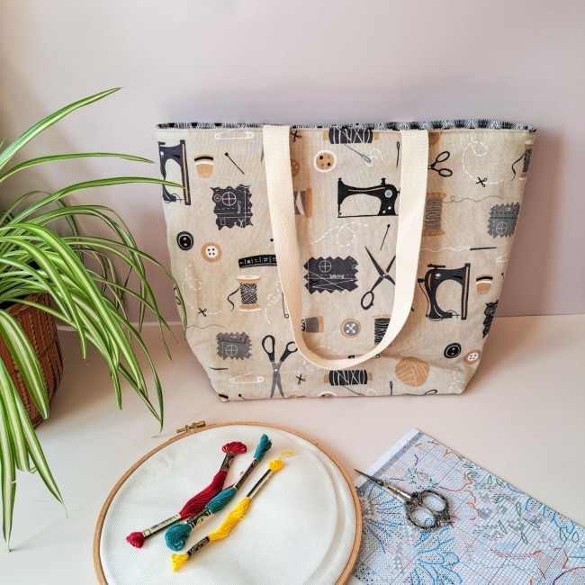 sac-cabas-rangement-broderie-angelique-la-fee-en-fil Posé sur la table près d'une plante, un sac à ouvrage Louise pour point de croix. de couleur beige avec des motifs de machibne à coudre. Devant un tambour à broder, des échevettes de fil à broder, un ciseau à broder et une grille de point de croix compté.