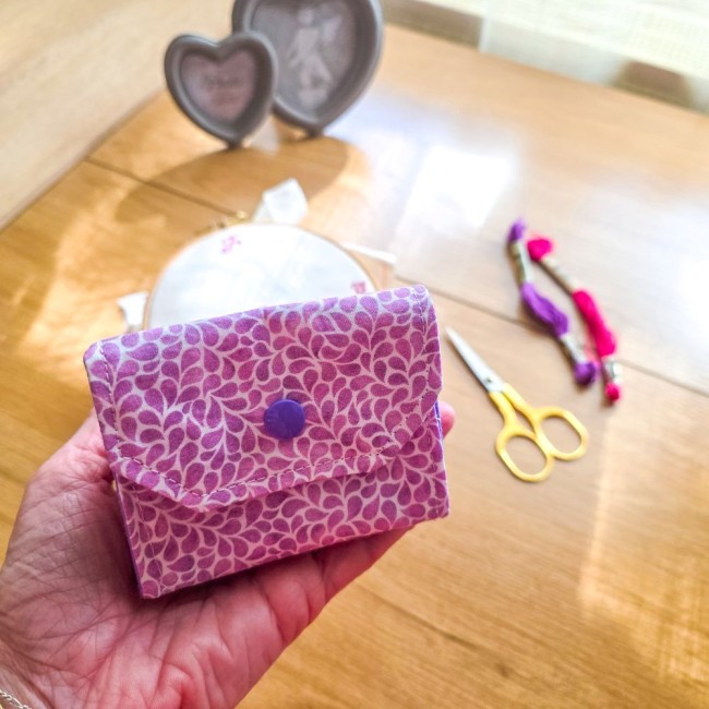 Vue de face en prise en main de la mini poubelle à fils de table pliante point de croix fermée modèle Lavande en tissu coton violet à motifs feuillage avec bouton-pression violet, posée au-dessus d'une table avec ciseaux dorés et échevettes, création arti