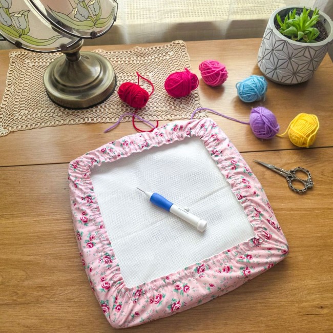 grime-guard-punch-needle-cadre-carre-romane-angelique-la-fee-en-fil Vue d'ensemble d'un grime guard housse protège-toile modèle Romane installé sur un cadre carré pour punch needle, en tissu rose aux motifs bucoliques de fleurs et roses, avec aiguille à puncher, pelotes de laine colorées, ciseaux vintage et lampe rétro su