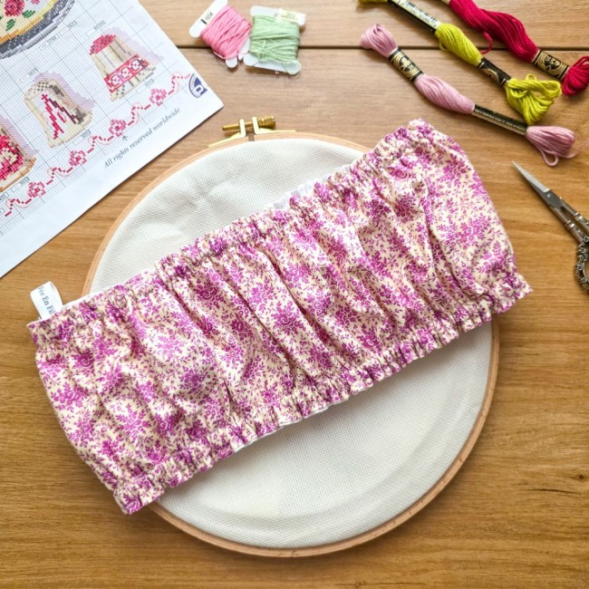 Vue d'ensemble d'un grime guard housse protège-toile modèle Samba en tissu coton à motifs floraux rose et violet, ajusté sur un tambour à broder rond en bois , avec l'étiquette de la marque visible, grille de broderie, échevettes de fils et ciseaux vintag