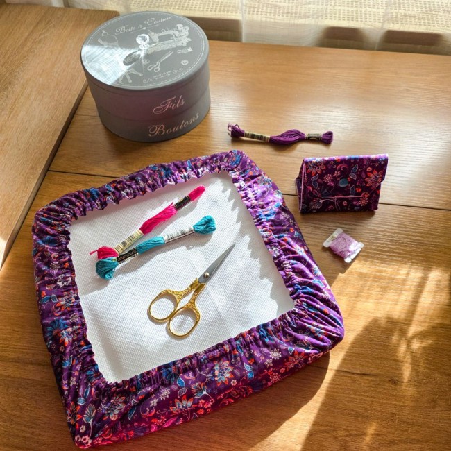 Vue de dessus d'un grime guard modèle Cachemire en tissu violet aux motifs floraux et cachemire multicolores, installé sur un cadre q-snap carré avec sa poubelle à fil assorti, des échevettes de fils, ciseaux dorés et une boîte à couture, création artisan
