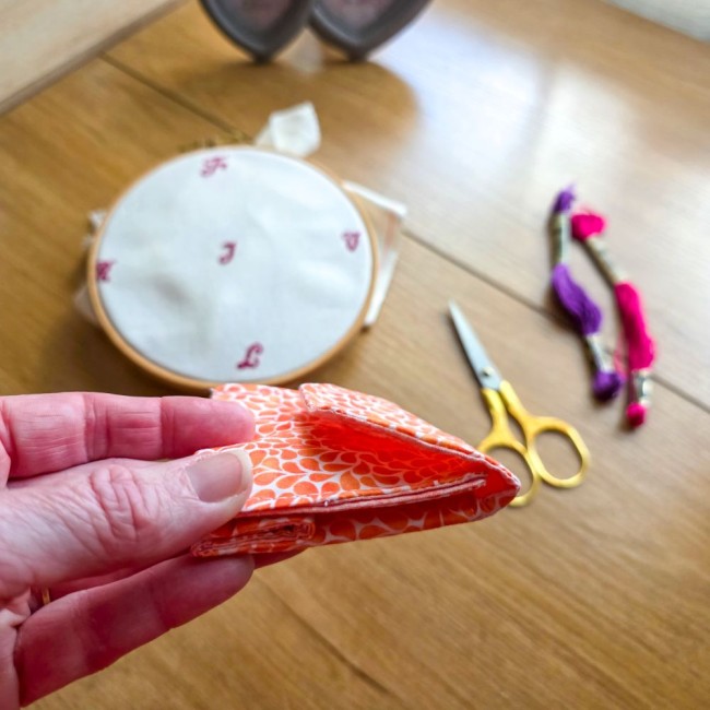 Mini poubelle à fils de table pliante modèle Mandarine en tissu coton orange à motifs gouttelettes blanches montrée repliée à plat dans la main pour illustrer son format compact, avec un tambour à broder, des ciseaux dorés et des échevettes en arrière-pla