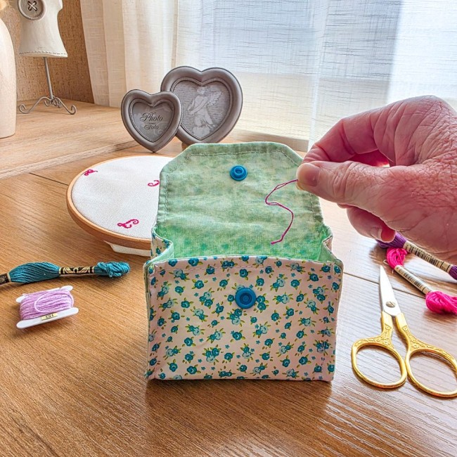 poubelle-a-fils-mini-pochon-broderie-myosotis-ouverte-utilisation-angelique-la-fee-en-fil Mini poubelle à fils pliable ouverte en tissu imprimé myosotis bleus et doublure verte, montrant une main y déposant un brin de fil rose, posée sur une table en bois avec du matériel de broderie, création artisanale Angélique La Fée En Fil.