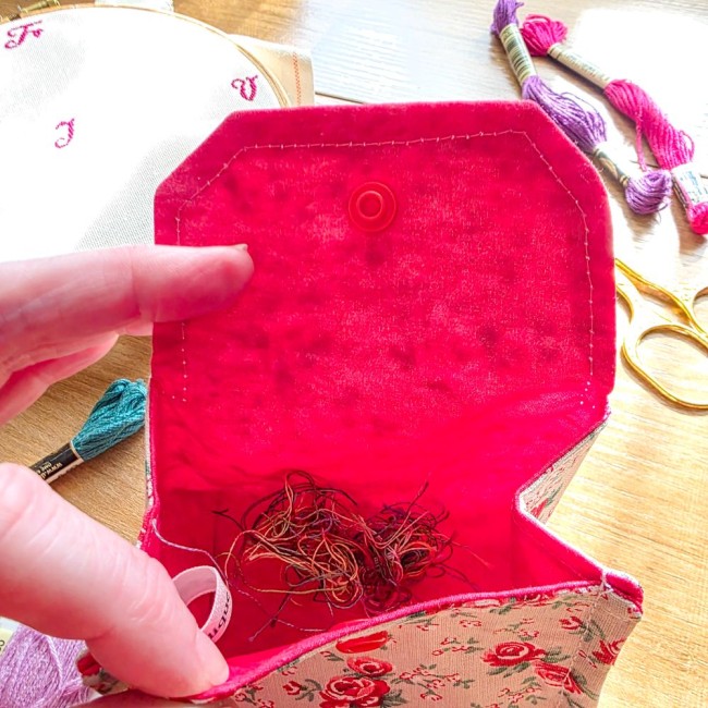 poubelle-a-fils-mini-pochon-broderie-vanille-interieur-dechets-fils-angelique-la-fee-en-fil Vue plongeante de l'intérieur d'une mini poubelle à fils ouverte modèle Vanille, montrant les chutes de fils de broderie au fond du pochon rouge fuchsia uni avec étiquette de marque, entourée de ciseaux dorés et d'échevettes, création artisanale Angélique