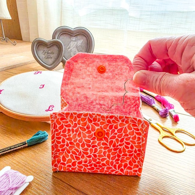 poubelle-a-fils-de-table-tricot-utilisation-mandarine-orange-angelique-la-fee-en-fil Mini poubelle à fils de table pliante modèle Mandarine en tissu coton orange à motifs gouttelettes blanches montrée en cours d'utilisation avec une main y jetant un fil de broderie, accompagnée d'un tambour, de ciseaux et d'échevettes, création artisanale