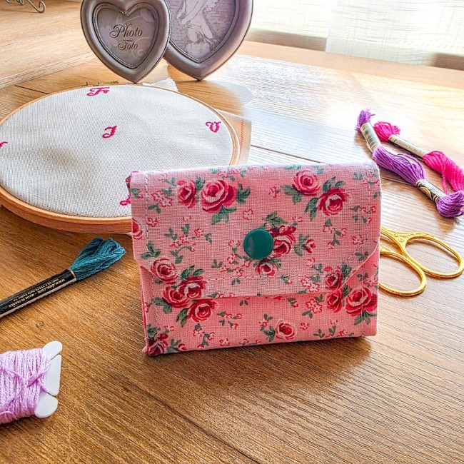Mini poubelle à fils pliable fermée modèle Romane en tissu rose imprimé de petites roses rouges avec bouton-pression vert, posée sur une table en bois avec du matériel de broderie, création artisanale Angélique La Fée En Fil.
