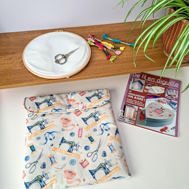 Mise en scène créative comprenant une pochette de rangement molletonnée aux motifs de machines à coudre anciennes, accompagnée d'un tambour à broder, de fils colorés, de ciseaux de brodeuse et d'un magazine 