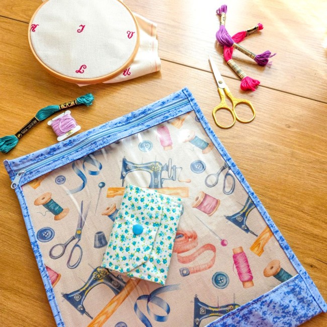 poubelle-a-fils-mini-pochon-myosotis-pochette-projet-broderie-angelique-la-fee-en-fil Mini poubelle à fils pliable imprimée de fleurs de myosotis bleues pliée à plat sur une pochette à projet transparente motif couture avec fermeture éclair bleue, entourée d'un tambour, ciseaux dorés et échevettes, création artisanale Angélique La Fée En F