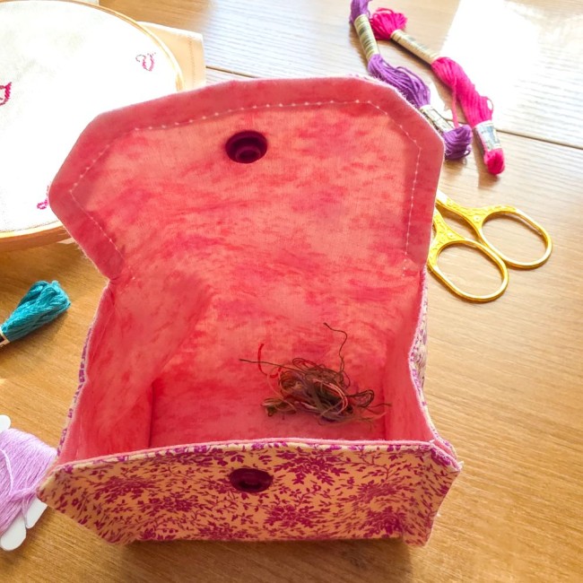 Vue en plongée d'une mini poubelle à fils de table ouverte modèle Samba en tissu coton à motifs floraux rose et violet, contenant quelques restes de fils coupés à l'intérieur, avec des ciseaux dorés et des échevettes sur le côté, création artisanale Angél