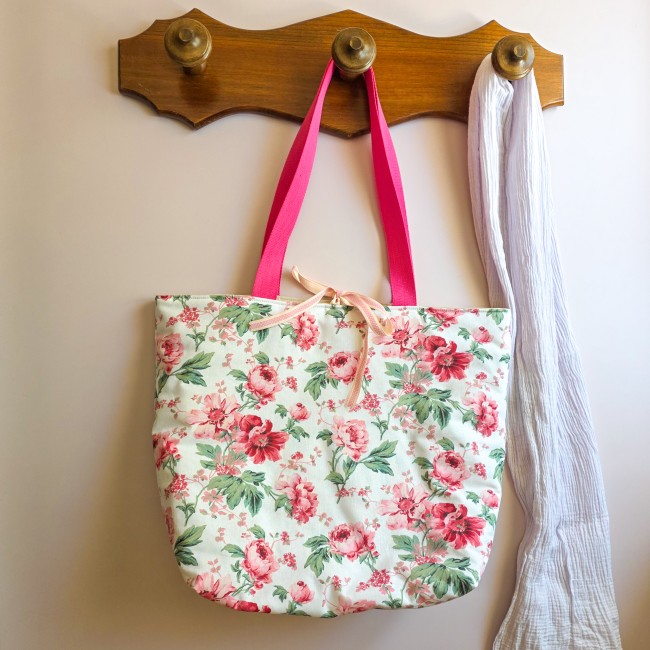 sac-cabas-sofia-tissu-fleurs-roses-anses-rose-angelique-la-fee-en-fil Sac cabas Sofia en tissu imprimé de fleurs roses et feuillage vert sur fond clair, suspendu à un porte-manteau en bois avec des anses rose vif.
