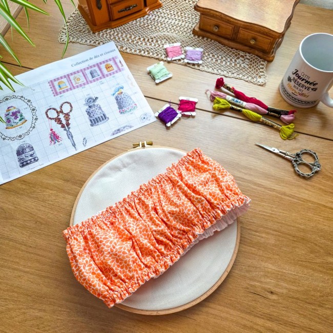 grime-guard-tambour-rond-mandarine-orange-angelique-la-fee-en-fil Vue d'ensemble d'un grime guard housse protège-toile modèle Mandarine en tissu coton orange imprimé de motifs gouttelettes blanches, ajusté sur un tambour à broder rond en bois, avec grille de broderie, ciseaux, échevettes de fils et mug sur une table en