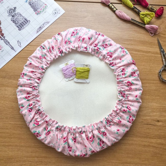grime-guard-tambour-rond-protege-toile-broderie-romane-angelique-la-fee-en-fil Vue de dessus d'un grime guard rond housse protège-toile modèle Romane installé sur un tambour à broder rond, en tissu rose aux motifs bucoliques de fleurs et roses, avec cartes de fils, ciseaux ciselés, échevettes et grille de point de croix sur une tabl