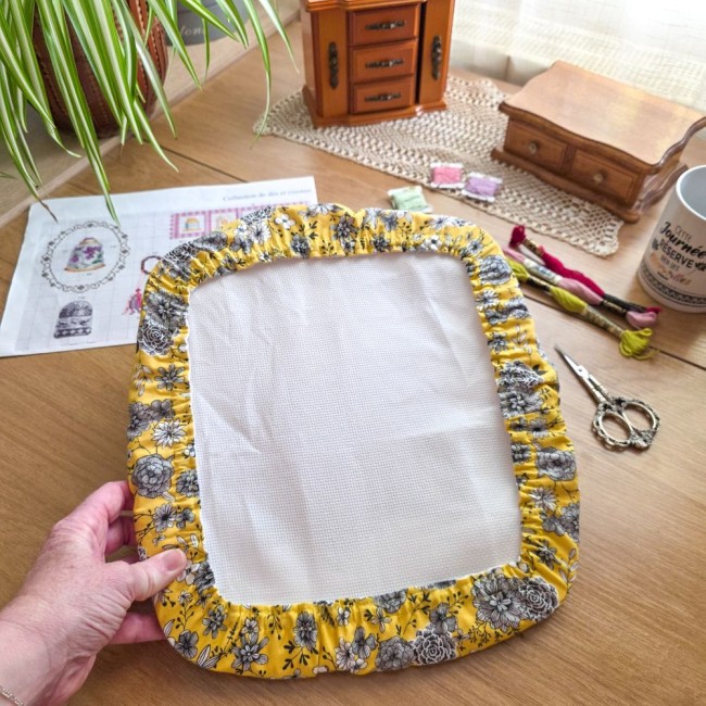 Vue de près d'une main tenant le grime guard modèle Julie en tissu jaune floral installé sur un métier Q-Snap, au-dessus d'une table avec accessoires de broderie par Angélique La Fée en Fil.