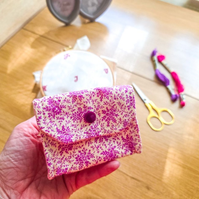 Mini poubelle à fils de table pliante modèle Samba en tissu coton à motifs floraux rose et violet présentée en prise en main, avec un tambour à broder, des ciseaux dorés et des échevettes en arrière-plan, création artisanale Angélique La Fée En Fil