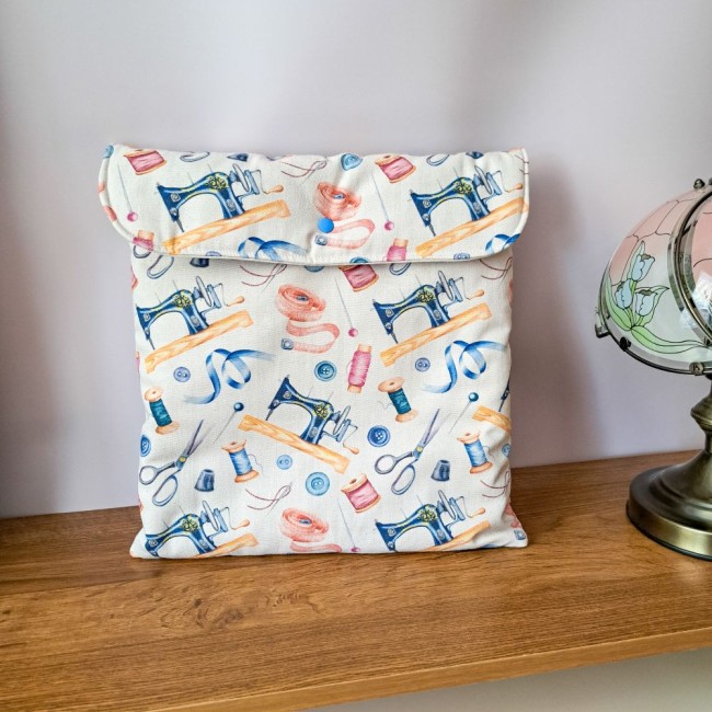Pochette de rangement pour tambour à broder posée verticalement sur une étagère en bois. Le tissu aux motifs rétro illustre des machines à coudre, des mètres rubans roses et des bobines de fil bleues. Une lampe de style est visible sur la droite. Création