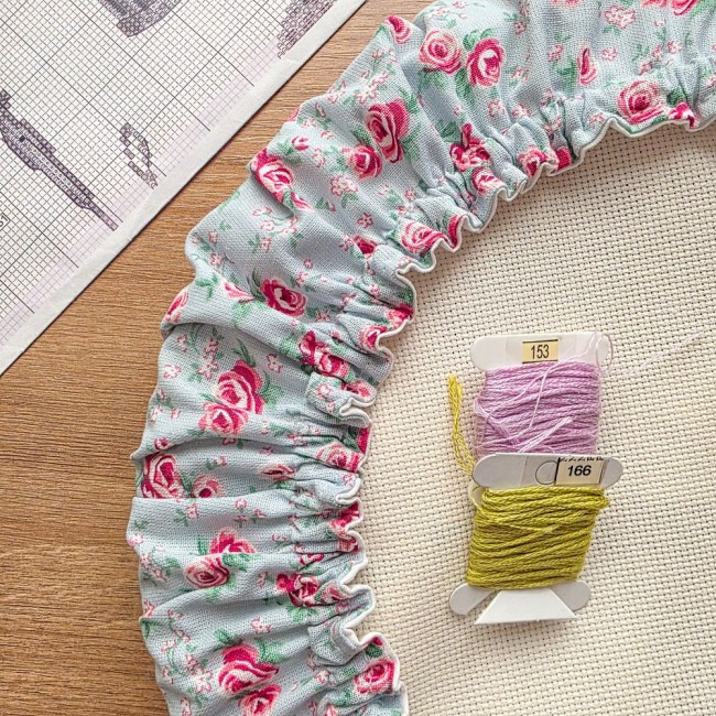 Gros plan sur les fronces et le tissu d'un grime guard modèle Bianca en tissu gris clair aux motifs bucoliques de roses rouges, posé sur une toile Aïda avec cartes de fils et grille de broderie en arrière-plan, création artisanale Angélique La Fée En Fil.