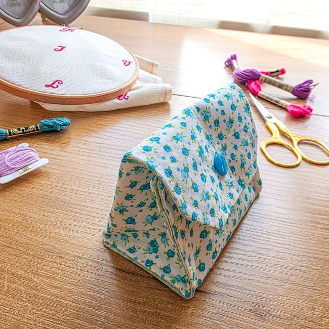 poubelle-a-fils-mini-pochon-broderie-myosotis-vue-profil-angelique-la-fee-en-fil Vue de profil et trois-quarts d'une mini poubelle à fils pliable ouverte en tissu écru imprimé de fleurs de myosotis bleues avec bouton-pression turquoise, posée sur une table en bois avec matériel de broderie, création artisanale Angélique La Fée En Fil.