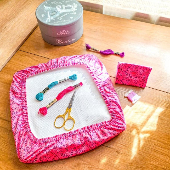 Ensemble d'accessoires de broderie assortis modèle Sandy Rose comprenant un grime guard sur cadre à broder et une mini poubelle à fils pliée en tissu rose aux motifs de feuillage violet, disposés sur une table en bois avec ciseaux dorés et échevettes, cré