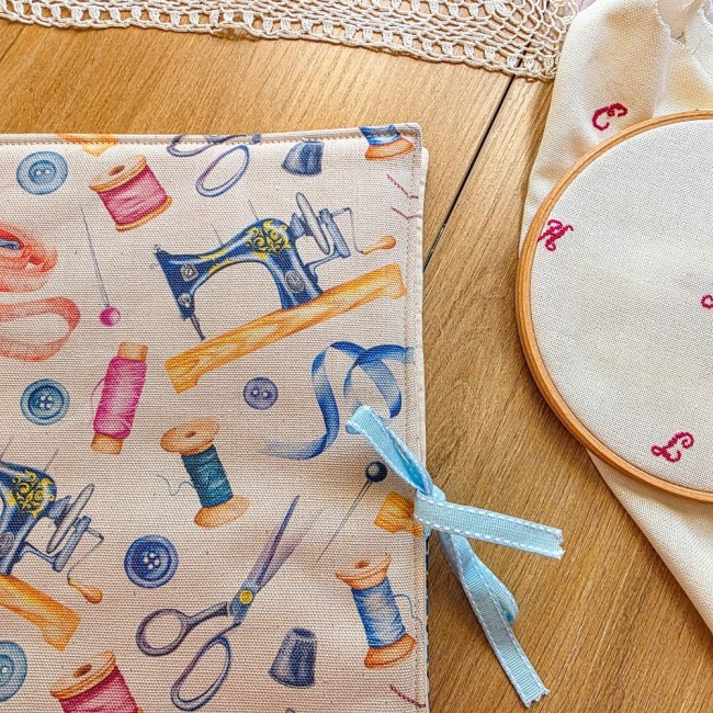 trousse-brodeuse-mercerie-vintage-mains-et-merveilles-angelique-la-fee-en-fil Gros plan sur l'extérieur de la trousse de brodeuse Mains et Merveilles en tissu aux motifs mercerie vintage, avec machine à coudre, ciseaux et bobines, fermée par un ruban bleu ciel, création artisanale Angélique La Fée En Fil.