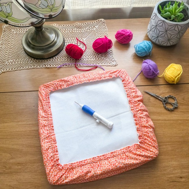 grime-guard-punch-needle-mandarine-orange-angelique-la-fee-en-fil Vue d'ensemble d'un grime guard housse protège-toile modèle Mandarine en tissu coton orange imprimé de motifs gouttelettes blanches, installé sur un support de broderie avec une aiguille magique pour punch needle, des pelotes de laine colorées, des ciseau