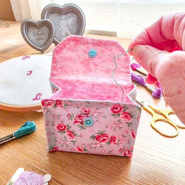 Gros plan d'une mini poubelle à fils ouverte en utilisation modèle Bianca, montrant l'intérieur en tissu rose clair avec dépôt d'un brin de fil de broderie, extérieure en tissu gris clair aux motifs bucoliques de roses rouges, création artisanale Angéliqu