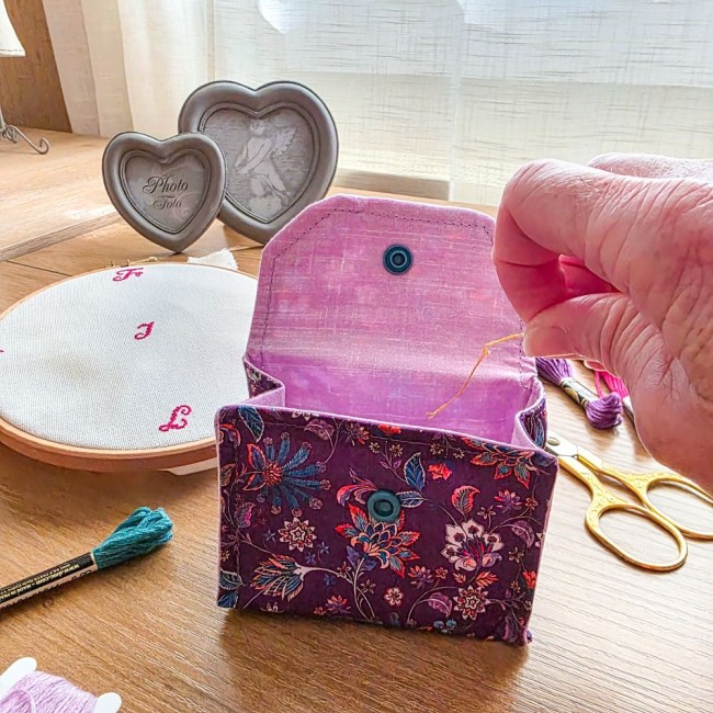 Mise en situation d'une mini poubelle à fils de table ouverte modèle Cachemire en tissu violet aux motifs floraux et cachemire multicolores, avec rabat rose, fil à broder déposé à l'intérieur, ciseaux dorés et tambour à broder en arrière-plan, création ar