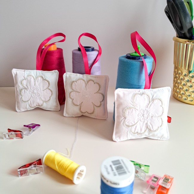 bijou-ciseaux-brodeuse-coussinet-fleur-blanche-ruban-rose-fils-bobines-couture-atelier-angelique-la-fee-en-fil Trois bijoux de ciseaux pour brodeuse en forme de coussinets blancs brodés d’une fleur, suspendus par des rubans roses, posés devant des bobines de fil rouge, violet et bleu, avec pinces de couture et bobines de fil sur une table d’atelier, création artis