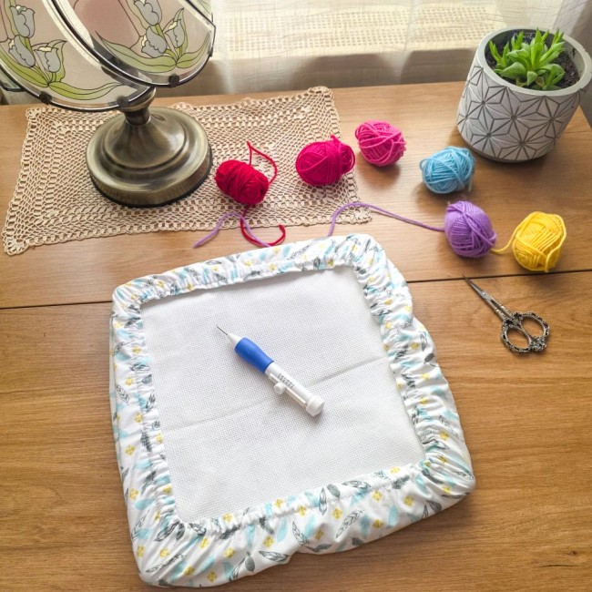 grime-guard-punch-needle-blanc-plumes-pastel-angelique-la-fee-en-fil Un grime guard (couvre-cadre) rectangulaire en coton blanc imprimé de plumes pastel bleues et grises et de petits losanges jaunes, protégeant une toile Aïda. Une aiguille magique à punch needle bleue et blanche est posée au centre. Décoration créative sur