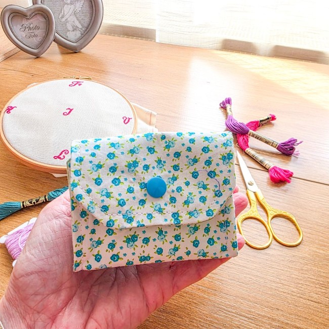 poubelle-a-fils-mini-pochon-broderie-myosotis-tenu-en-main-angelique-la-fee-en-fil Mini poubelle à fils pliable en tissu écru à motifs myosotis bleus tenue dans le creux de la main pour montrer sa taille compacte, avec ciseaux dorés et échevettes sur une table en bois, création artisanale Angélique La Fée En Fil.