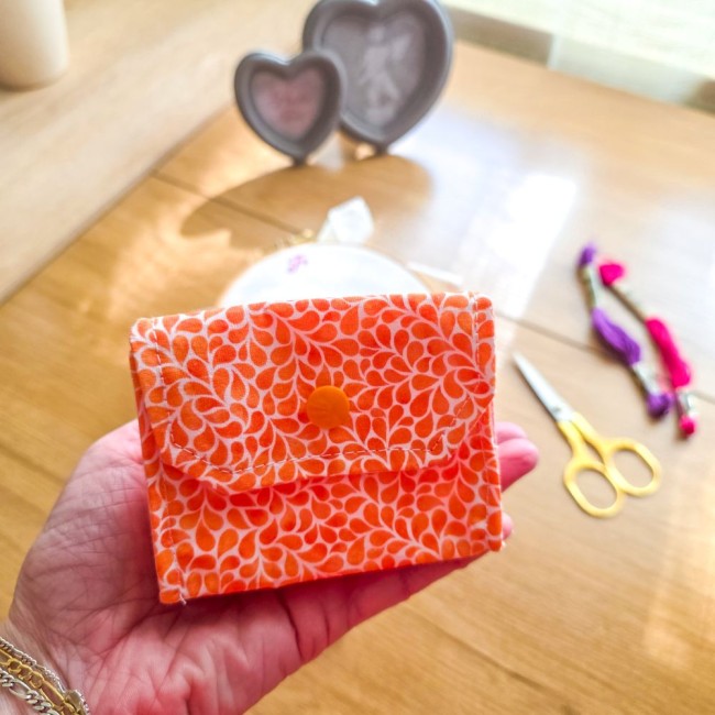 Mini poubelle à fils de table pliante modèle Mandarine en tissu coton orange à motifs gouttelettes blanches présentée en prise en main pour illustrer son format compact et pliable, avec des ciseaux dorés et un tambour à broder en arrière-plan, création ar