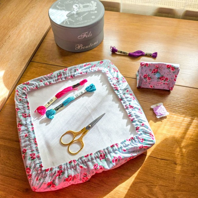 Ensemble d'accessoires de broderie assortis modèle Bianca comprenant un grime guard sur cadre à broder et une mini poubelle à fils pliée en tissu gris clair aux motifs bucoliques de roses rouges, disposés sur une table en bois avec ciseaux dorés et écheve