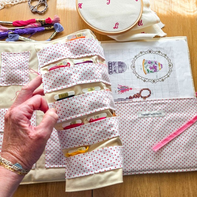 trousse-brodeuse-rangement-cartonnettes-grille-a-broder-alice-angelique-la-fee-en-fil Main soulevant le rabat de cartonnettes de la trousse de brodeuse Alice à pois rouges, dévoilant l'emplacement pour la grille de point de croix, création artisanale Angélique La Fée En Fil.