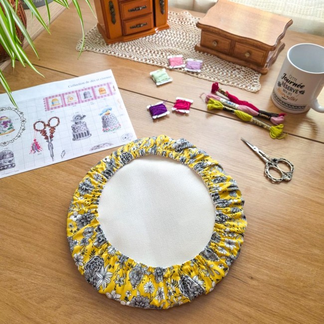 Grime guard rond modèle Julie en tissu jaune imprimé de fleurs noires, houssant un tambour à broder sur une table en bois entouré de fil et ciseaux par Angélique La Fée en Fil.