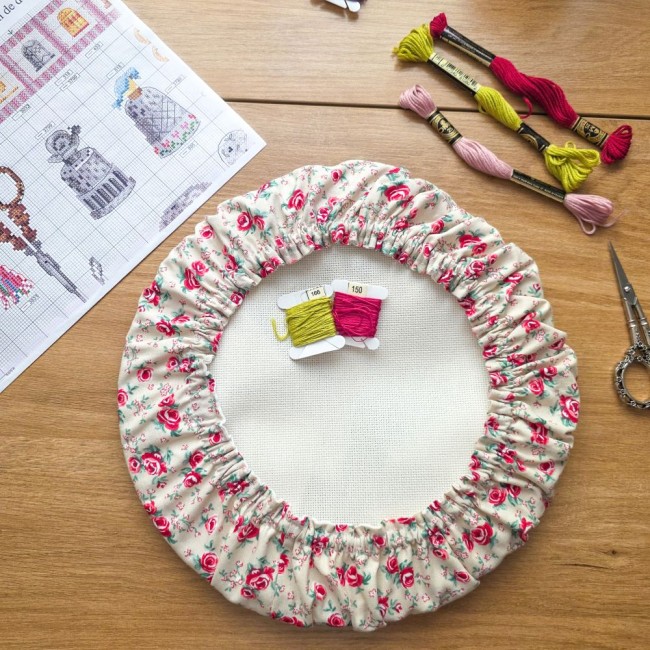 Vue de dessus d'un grime guard rond housse protège-toile modèle Vanille installé sur un tambour à broder rond, en tissu écru vanille aux motifs bucoliques de roses roses et rouges, avec cartes de fils, ciseaux ciselés, échevettes et grille de point de cro
