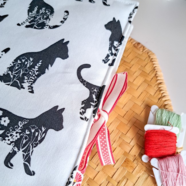 Trousse de brodeuse Jardin des Félins en tissu blanc imprimé de silhouettes de chats noirs fleuris, nouée d’un ruban rouge, création artisanale Angélique La fée En Fil, présentée avec fils à broder rouge et vert.