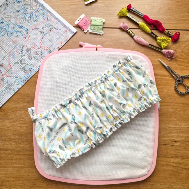 grime-guard-sur-cadre-nurge-rose-blanc-plumes-pastel-angelique-la-fee-en-fil Un couvre-cadre (grime guard) élastiqué en coton blanc imprimé de plumes pastel bleues et grises et de petits losanges jaunes, posé en diagonale sur une toile Aïda blanche tendue dans un cadre carré rose de marque Nurge. L'étiquette de la marque est visib