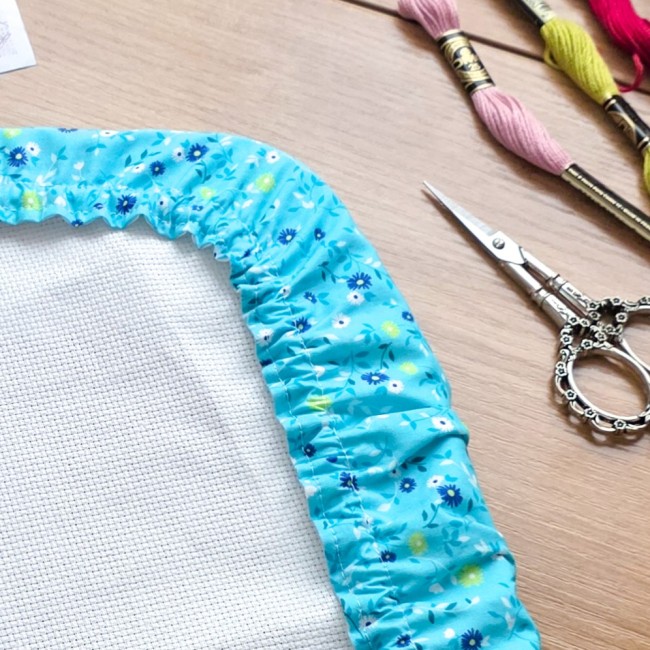 grime-guard-broderie-bleu-turquoise-fleuri-gros-plan-coin-angelique-la-fee-en-fil Gros plan sur l'angle d'un grime guard (couvre-cadre) en coton bleu turquoise imprimé de petites fleurs blanches et jaunes, ajusté sur un cadre avec sa toile Aïda blanche. On aperçoit la finition des coutures et de l'élastique ainsi qu'une paire de ciseau