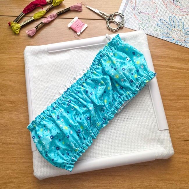 grime-guard-sur-cadre-qsnap-blanc-bleu-turquoise-fleuri-angelique-la-fee-en-fil Un couvre-cadre (grime guard) élastiqué en coton bleu turquoise imprimé de petites fleurs blanches, bleues et jaunes, posé en diagonale sur une toile Aïda blanche fixée sur un cadre rectangulaire blanc de type Q-Snap. L'étiquette de la marque est visible.