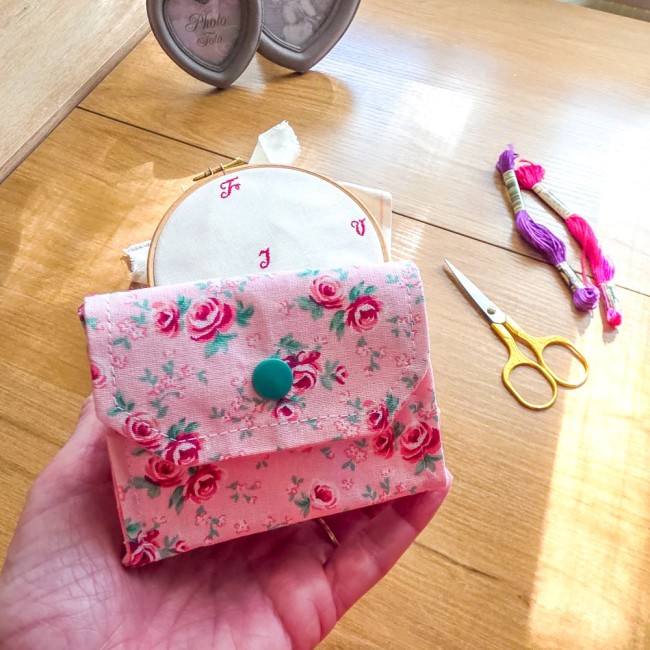 Mini poubelle à fils nomade tenue à plat dans la main fermée par un bouton-pression vert, modèle Romane en tissu rose à motif de roses rouges, avec tambour à broder, ciseaux dorés et échevettes de fils en arrière-plan, création artisanale Angélique La Fée