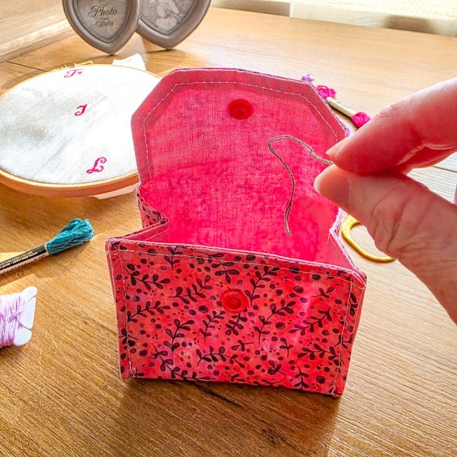 Gros plan d'une mini poubelle à fils ouverte en utilisation modèle Sandy Rose, montrant l'intérieur en tissu rose vif avec dépôt d'un brin de fil de broderie, extérieure en tissu rose aux motifs de feuillage violet, création artisanale Angélique La Fée En