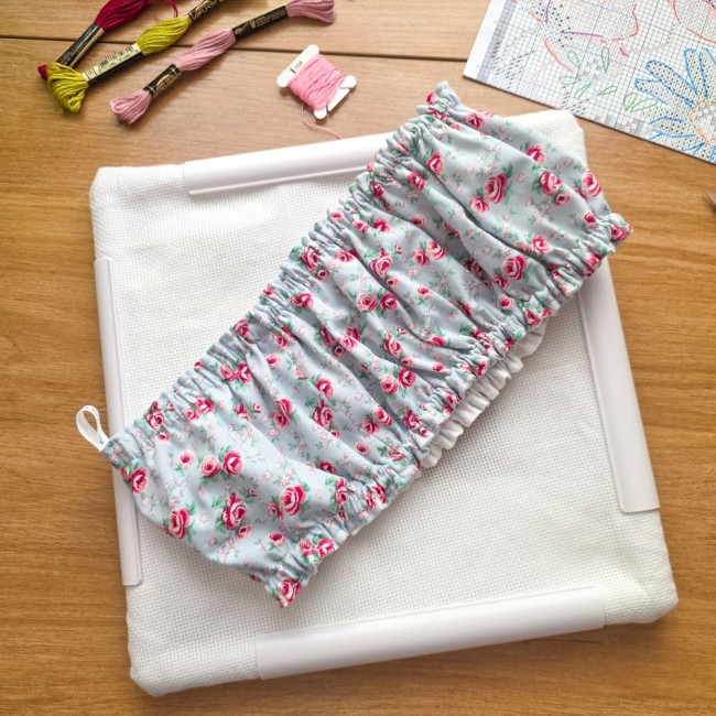Vue de dessus d'un grime guard housse protège-toile modèle Bianca posé en travers sur un cadre Q-Snap carré blanc avec toile Aïda, en tissu gris clair aux motifs bucoliques de roses rouges avec petite boucle d'accroche, entouré d'échevettes et d'une grill