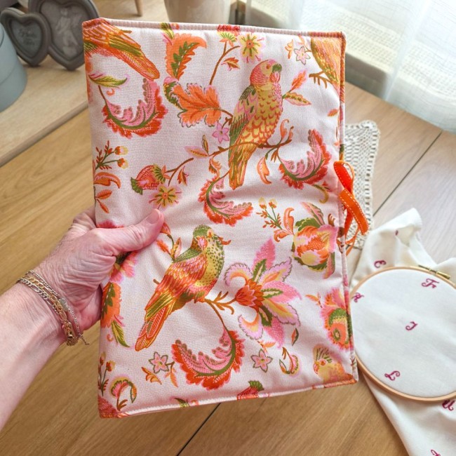 Main tenant la trousse de brodeuse Savannah fermée, montrant sa couverture en tissu imprimé d'oiseaux et de fleurs exotiques avec son lien en ruban orange, création artisanale Angélique La Fée En Fil.