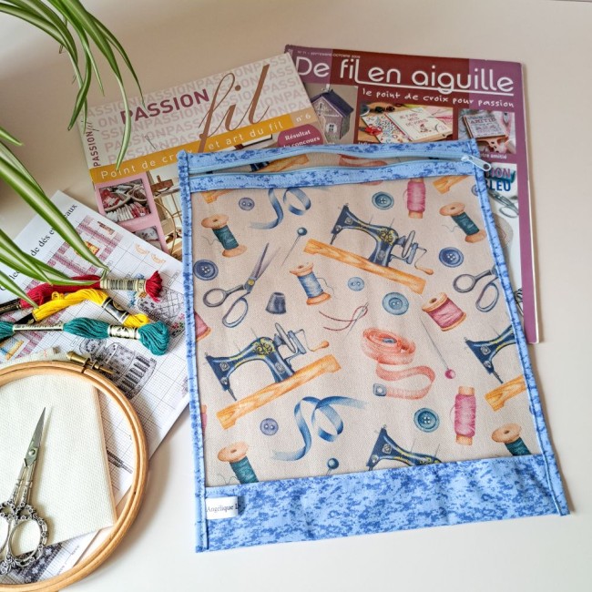  Posé sur la table près d'une plante, une pochette à projet de coulmeur bleu avec un motif de couture fermé par une fermeture éclair. A côté, des magazines de broderie, des echevettes, un ciseaux à broder, une grille de point compté et une toile à broder.