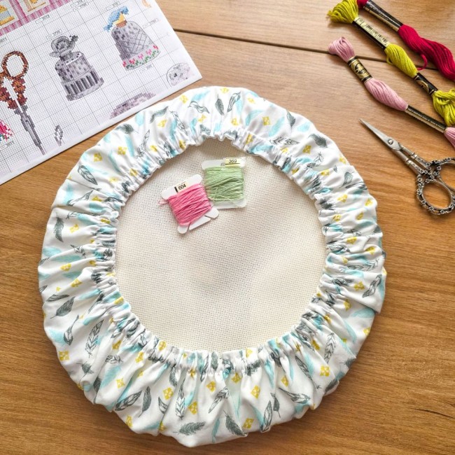 grime-guard-tambour-rond-blanc-plumes-pastel-point-de-croix-angelique-la-fee-en-fil Un couvre-tambour (grime guard) rond en coton blanc imprimé de plumes pastel bleues et grises et de petits losanges jaunes, installé sur un cercle à broder avec sa toile blanche. Deux cartonnettes de fils (rose n°604 et vert d'eau n°369) sont posées au ce
